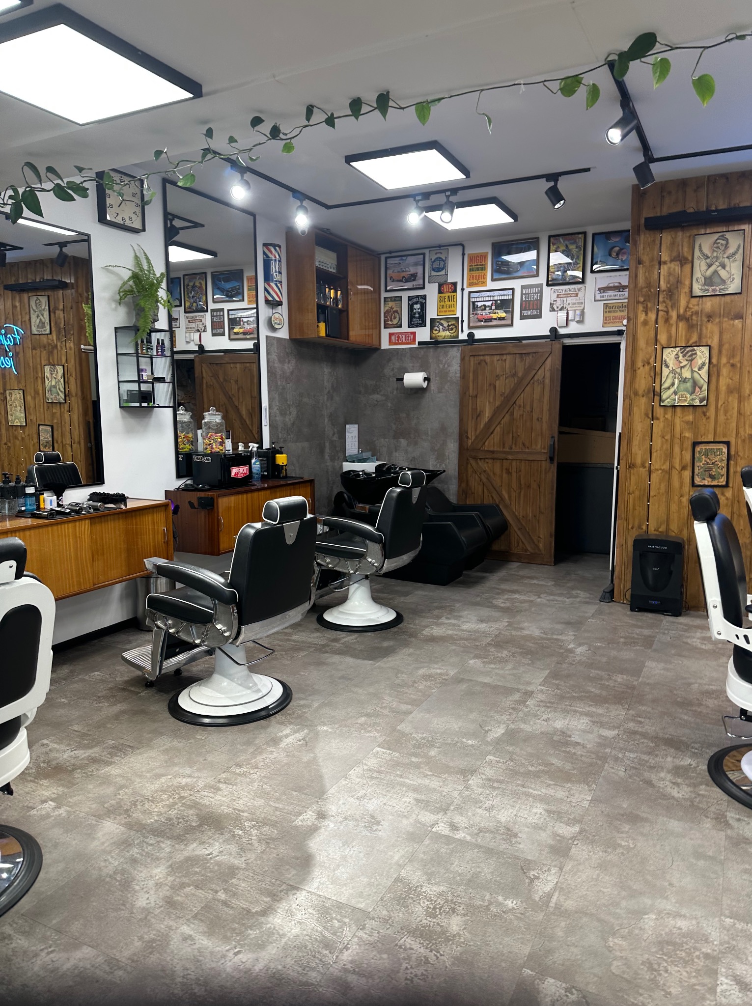 Nowoczesny wystrój salonu barberskiego: czarne fotele barberskie, drewniane akcenty, plakaty vintage i rośliny pnące się po suficie. Widok na strefę mycia głowy.
