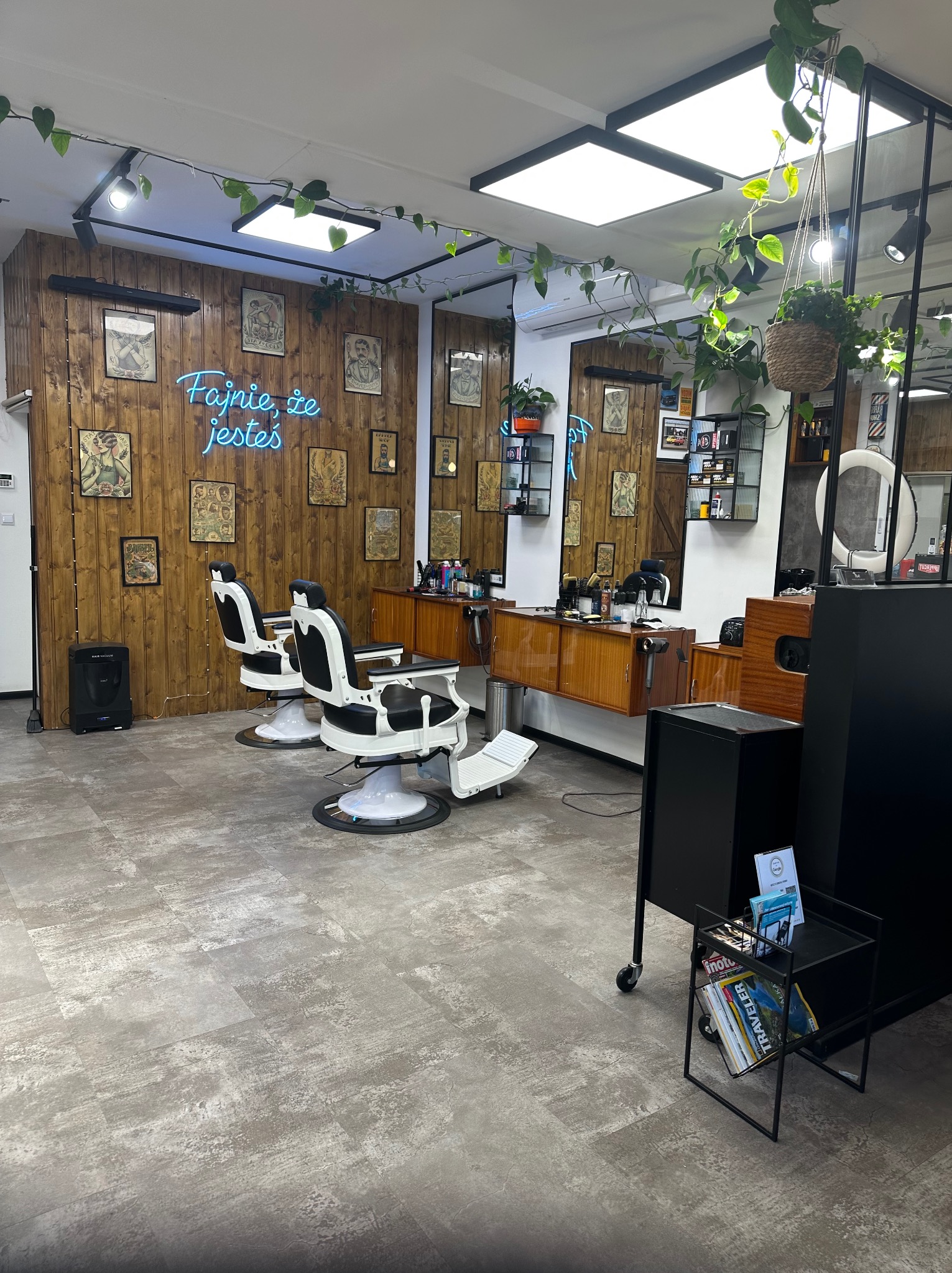 Wnętrze nowoczesnego salonu fryzjerskiego z drewnianymi panelami, neonem 'Fajnie, że jesteś', dwoma fotelami barberskimi i roślinami doniczkowymi.