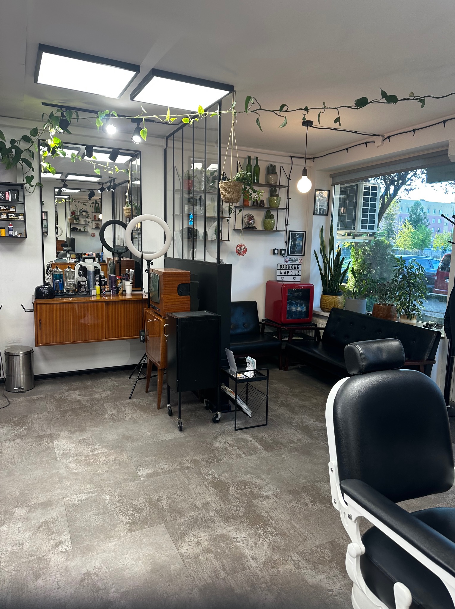 Wnętrze barbershopu w stylu retro: drewniane meble, rośliny pnące, czarne fotele, oświetlenie LED i dekoracyjny neon z napisem 'Darmowe napoje'.