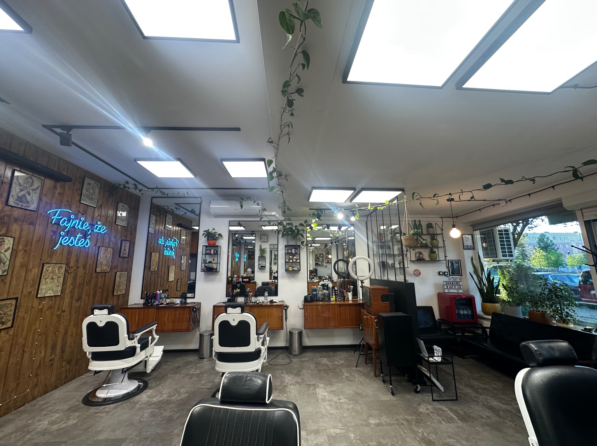 Wnętrze nowoczesnego salonu fryzjerskiego z drewnianą ścianą, neonem i roślinami. Widoczne stanowiska z fotelami fryzjerskimi i lustrami.