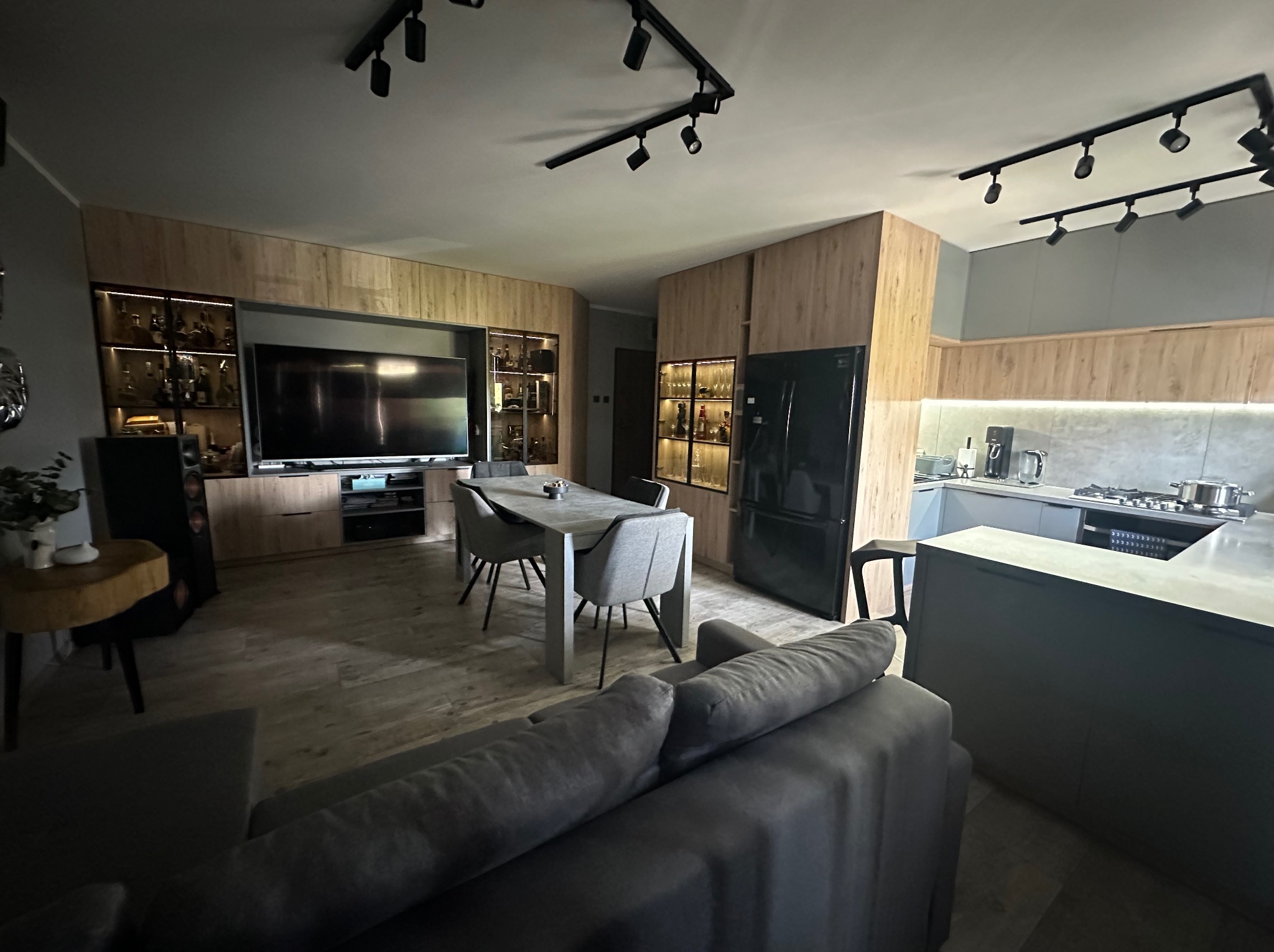 Nowoczesne wnętrze: salon z jadalnią i kuchnią w odcieniach szarości i drewna. Oświetlenie punktowe, minimalistyczny design, czarna lodówka i stół z szarymi krzesłami.