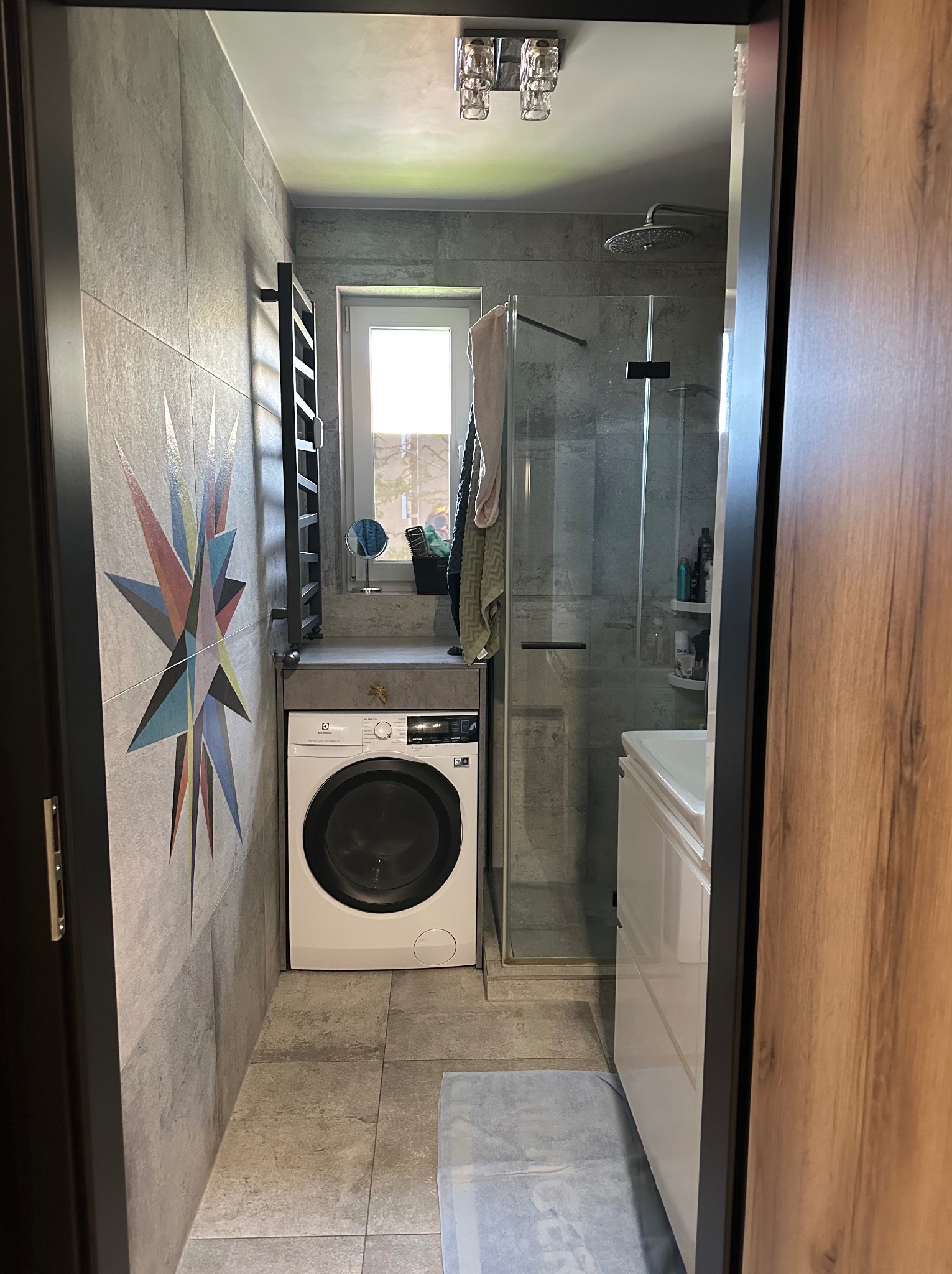 Szara łazienka z kabiną prysznicową i pralką Electrolux. Na ścianie dekoracja w kształcie gwiazdy. Nowoczesne wnętrze z szarymi płytkami.