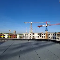 Nowe krycie dachu papą termozgrzewalną na tle budowy z żurawiami i kominem, w słoneczny dzień.