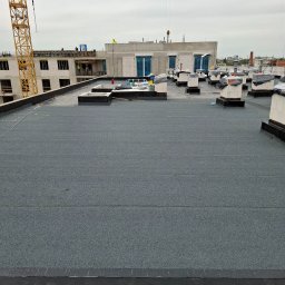 Fast Dariusz Pasternak - Szary, płaski dach budynku w budowie z kominami wentylacyjnymi, widok z góry. W tle żuraw budowlany i konstrukcja budynku z pracownikami.