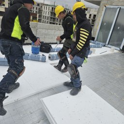 Fast Dariusz Pasternak - Praca dekarzy na dachu budynku w budowie. Widoczne żółte kaski, izolacja termiczna i rolki papy. W tle dźwig budowlany i budynki w trakcie wznoszenia.