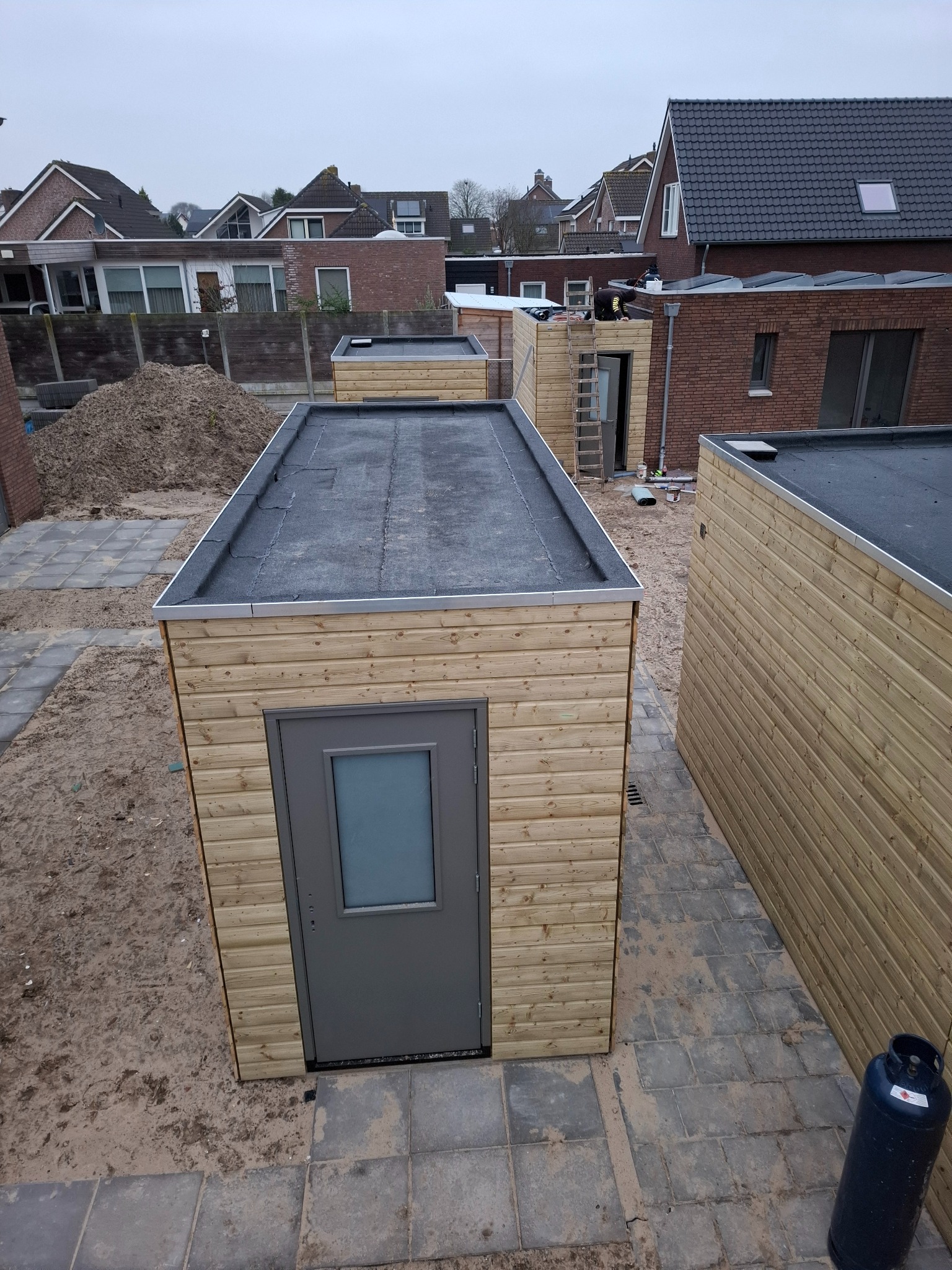 Montaż płaskiego dachu na drewnianych domkach gospodarczych w zabudowie szeregowej, widoczny dekarz na jednym z dachów i butla gazowa obok domku.