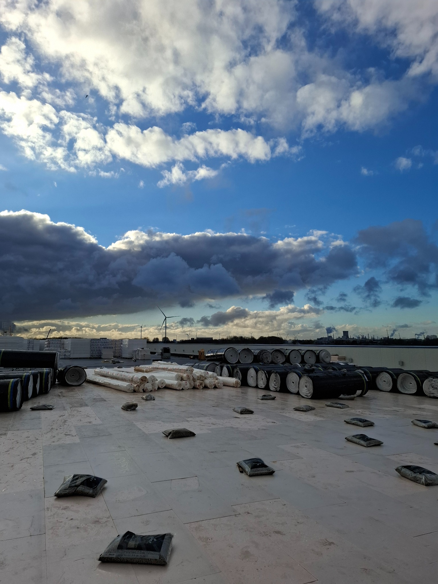 Materiały budowlane, w tym rolki papy i izolacji, rozłożone na płaskim dachu z widocznymi obciążnikami, na tle pochmurnego nieba z elementami krajobrazu przemysłowego i turbinami wiatrowymi w oddali.