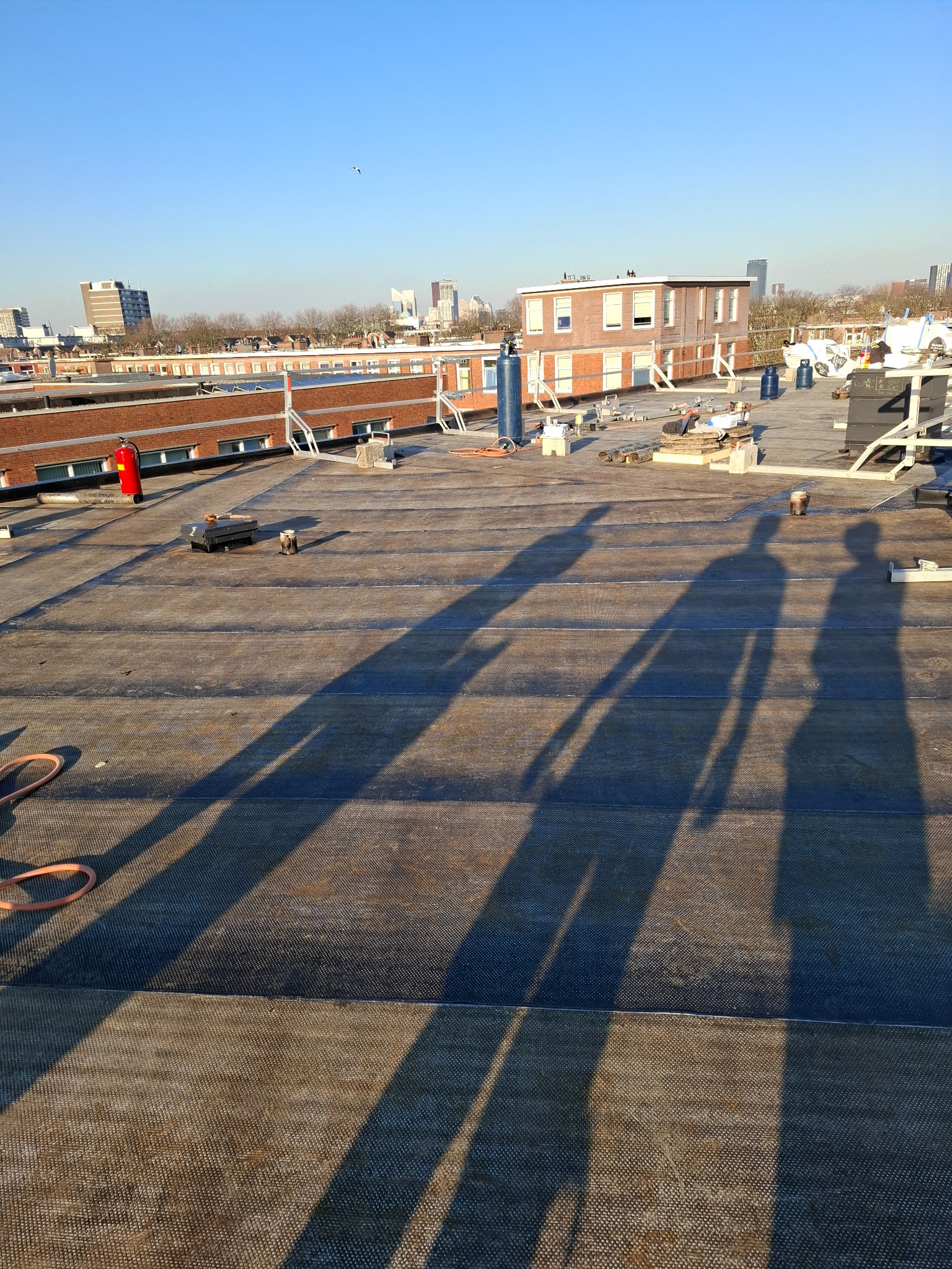 Rozległy płaski dach pokryty papą, widoczne narzędzia, butle z gazem, osoba w czerwonym ubraniu w oddali oraz długie cienie trzech osób na powierzchni dachu, w tle zabudowa miejska w słoneczny dzień.