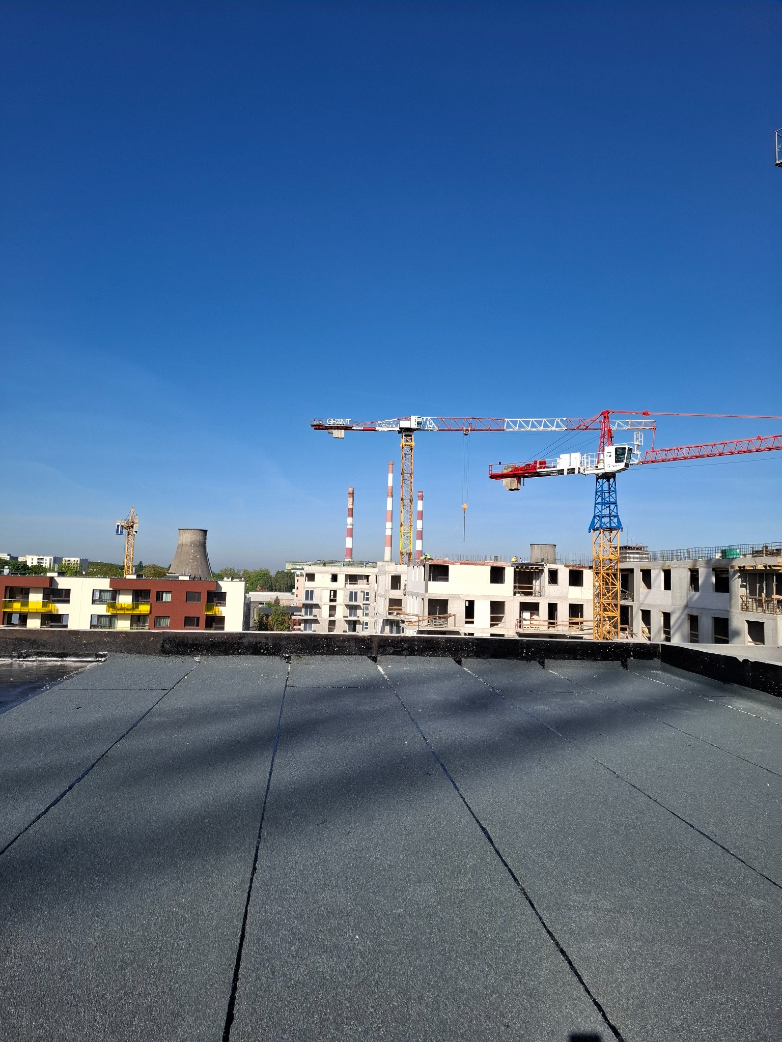 Nowe krycie dachu papą termozgrzewalną na tle budowy z żurawiami i kominem, w słoneczny dzień.