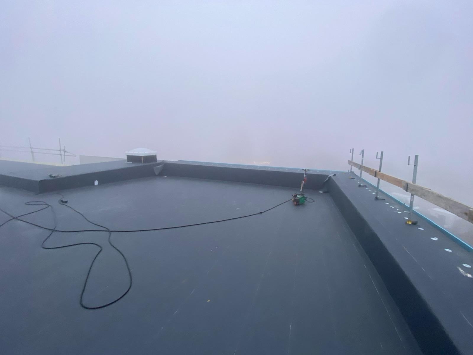 Szary, płaski dach pokryty membraną, z widocznym sprzętem do zgrzewania i elementami zabezpieczeń krawędzi, spowity gęstą mgłą.