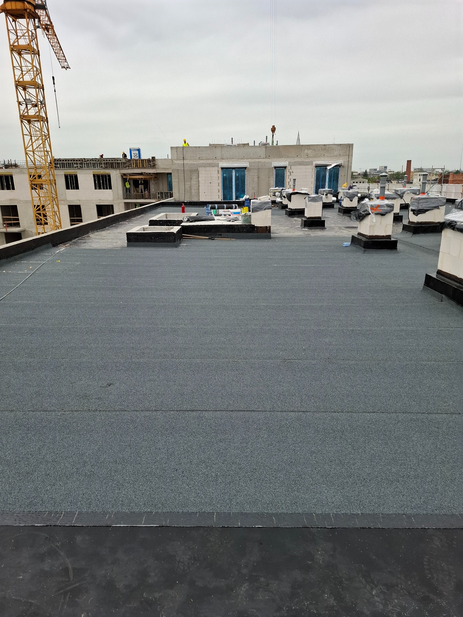 Szary, płaski dach budynku w budowie z kominami wentylacyjnymi, widok z góry. W tle żuraw budowlany i konstrukcja budynku z pracownikami.