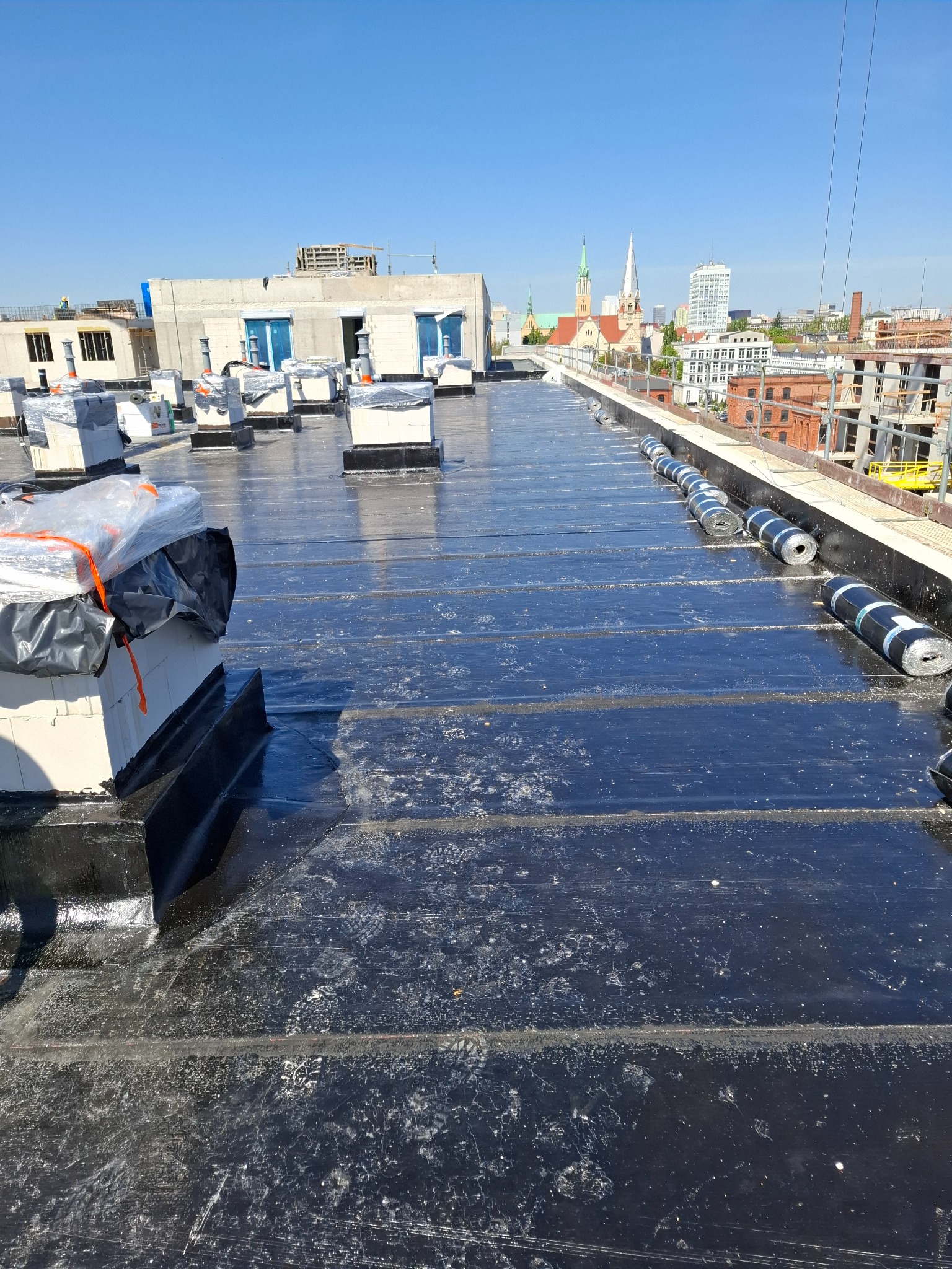 Wykończony dach pokryty czarną papą termozgrzewalną na budynku w mieście. Widoczne kominy wentylacyjne i rolki papy. W tle zabudowa miejska i kościół.