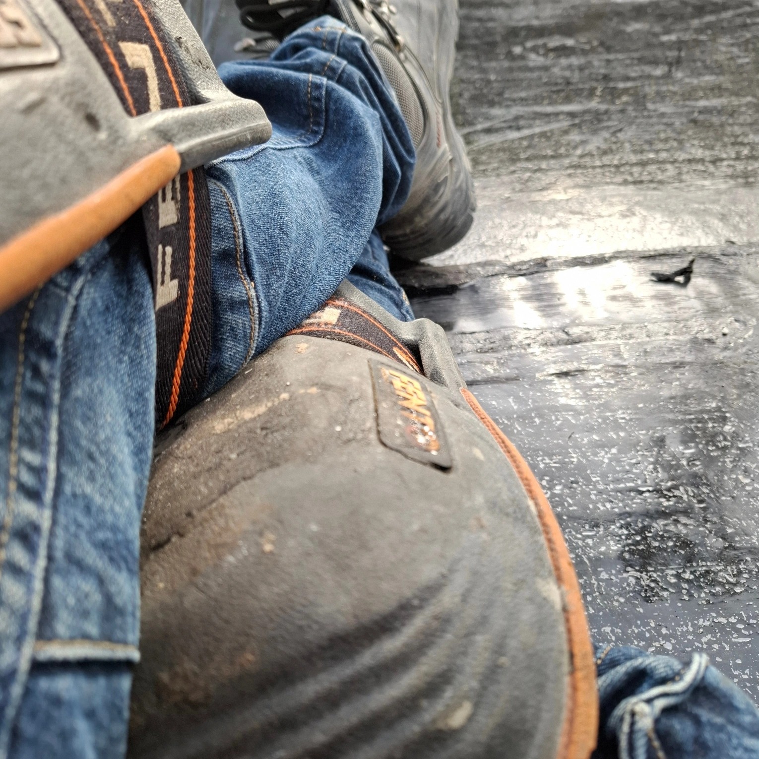 Pracownik w jeansach i nakolannikach siedzi na ciemnej, płaskiej powierzchni dachu. W tle widoczne buty robocze.