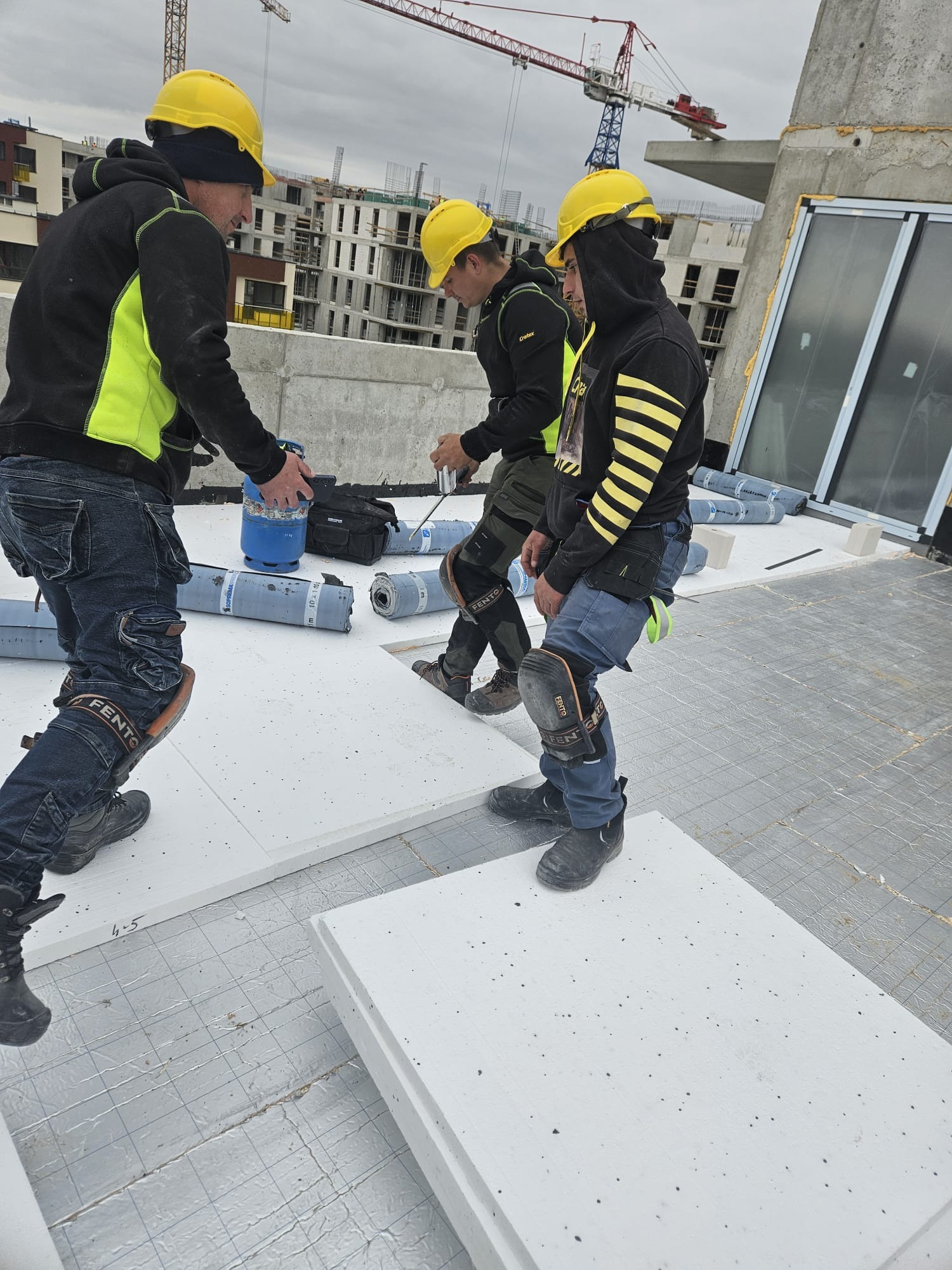 Praca dekarzy na dachu budynku w budowie. Widoczne żółte kaski, izolacja termiczna i rolki papy. W tle dźwig budowlany i budynki w trakcie wznoszenia.