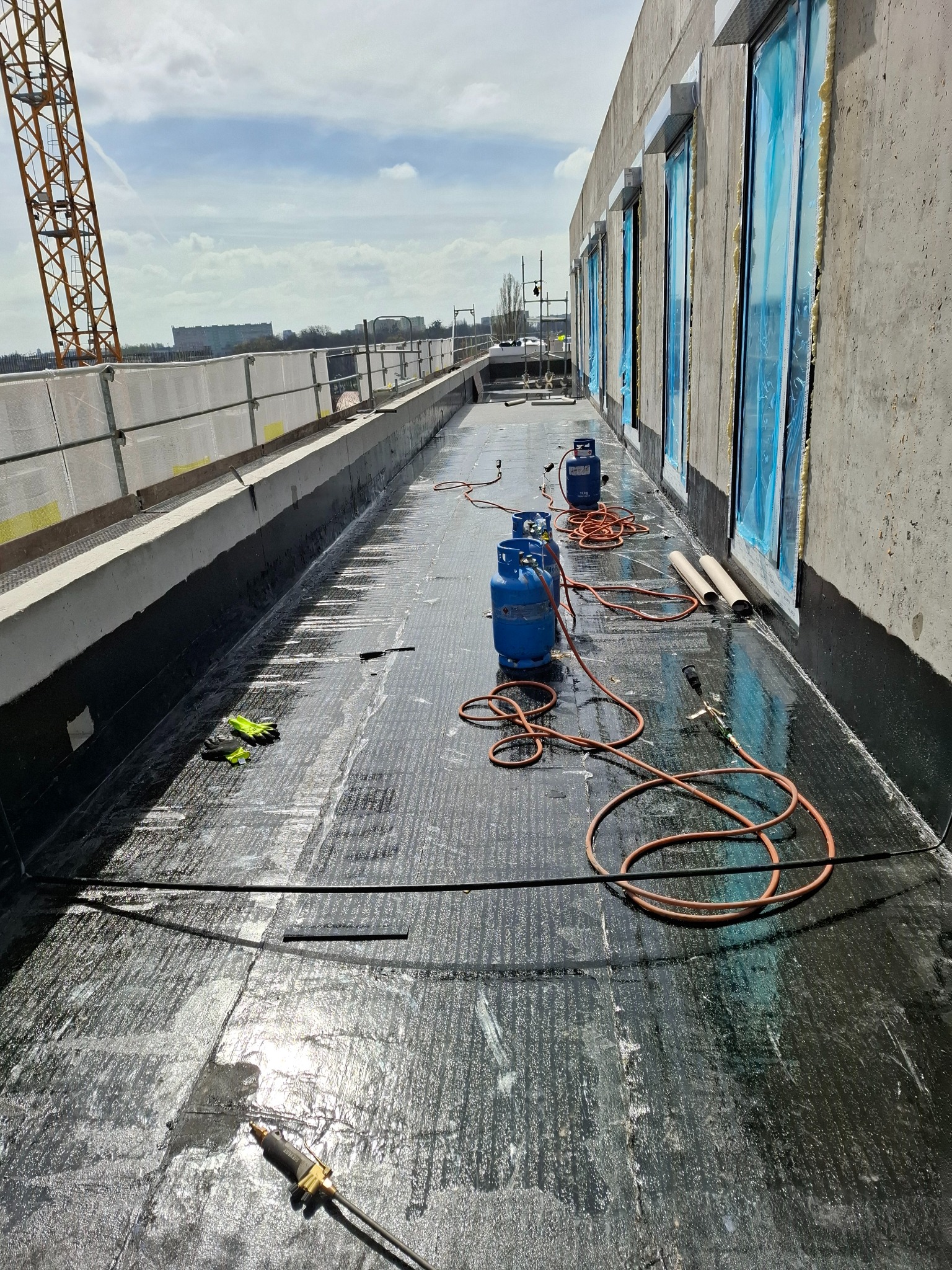 Czarna hydroizolacja dachu płaskiego w trakcie prac. Widoczne butle gazowe, palnik dekarski, rękawice robocze i zabezpieczone okna. W tle dźwig budowlany.