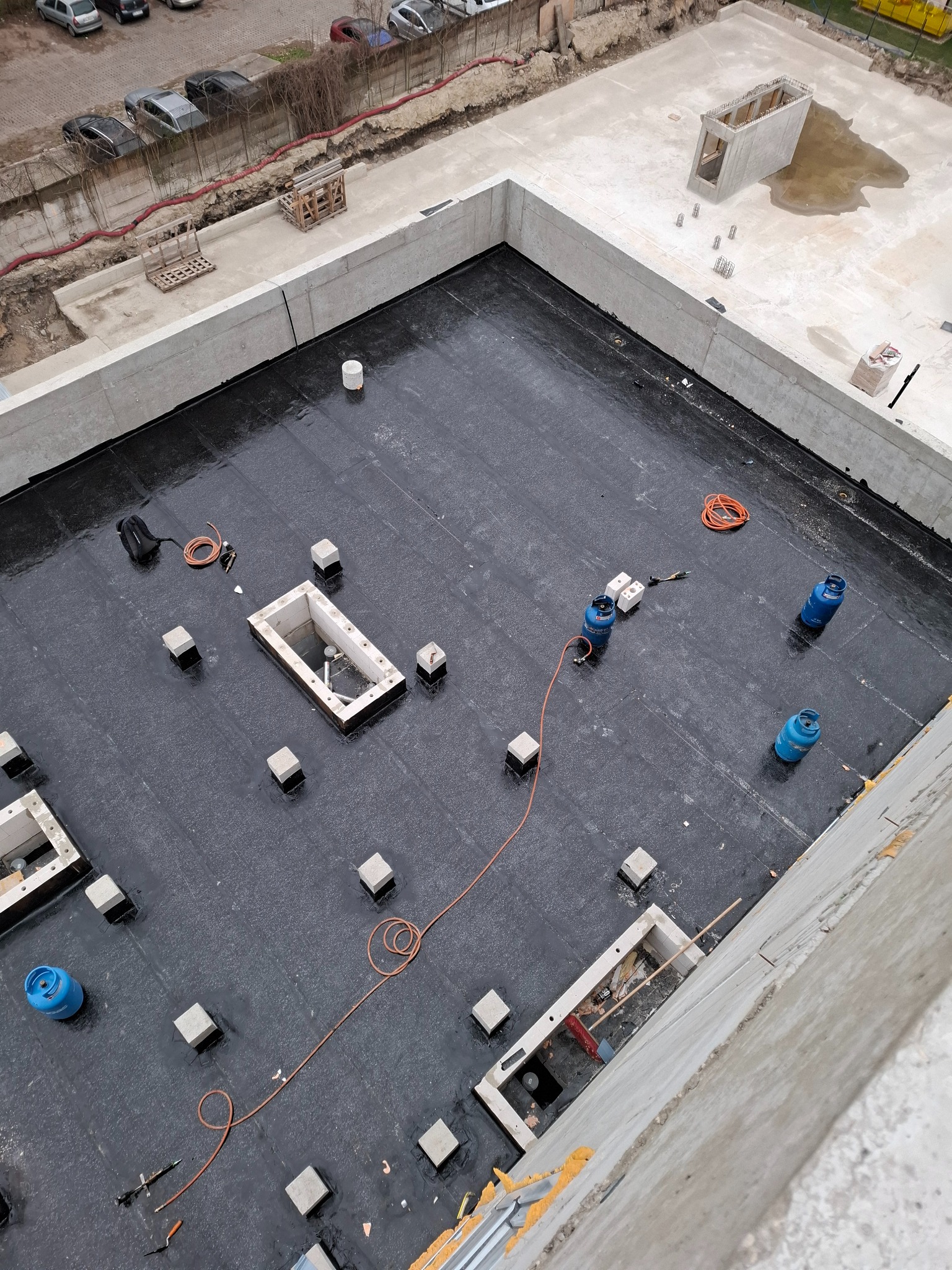 Widok z góry na czarną, płaską powierzchnię dachu z otworami wentylacyjnymi, betonowymi wspornikami, butlami gazowymi i narzędziami dekarskimi w trakcie prac.