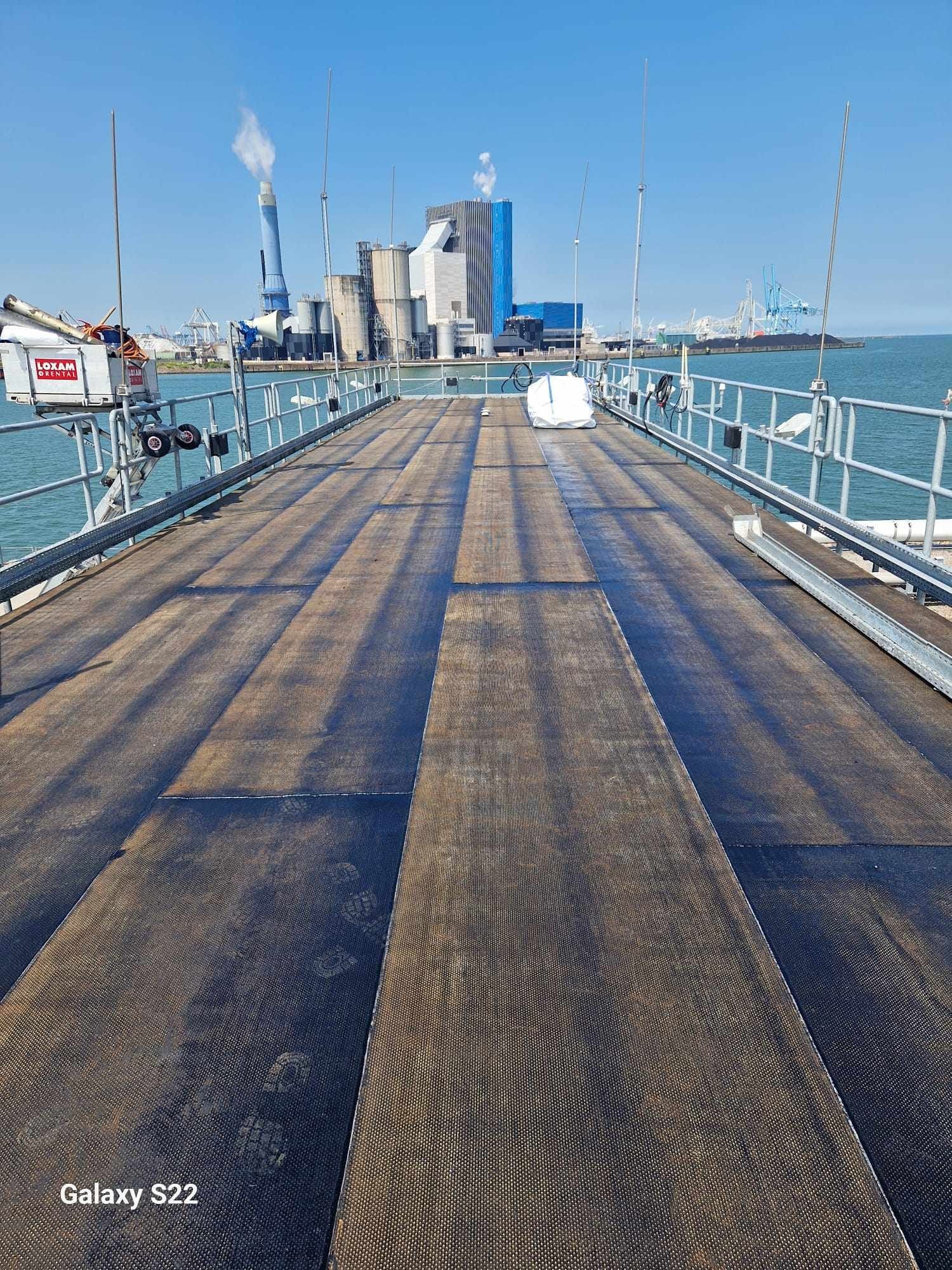 Nowa, płaska powierzchnia dachu przemysłowego z widocznymi śladami stóp, zabezpieczona barierkami. W tle zabudowania przemysłowe i zbiorniki.