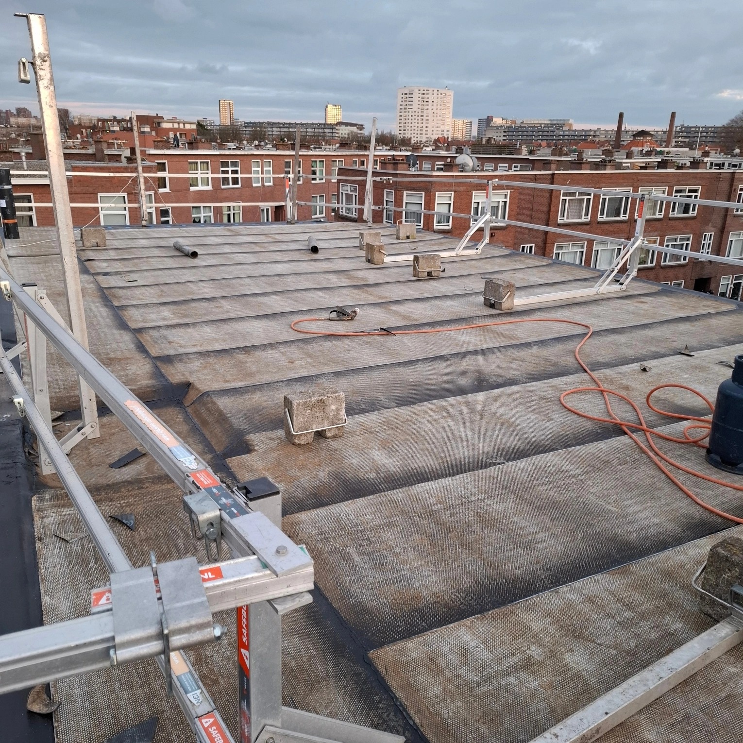 Płaski dach pokryty papą z widocznymi elementami zabezpieczeń, na tle zabudowy miejskiej w pochmurny dzień. Widoczne złączenia papy i ciężarki.