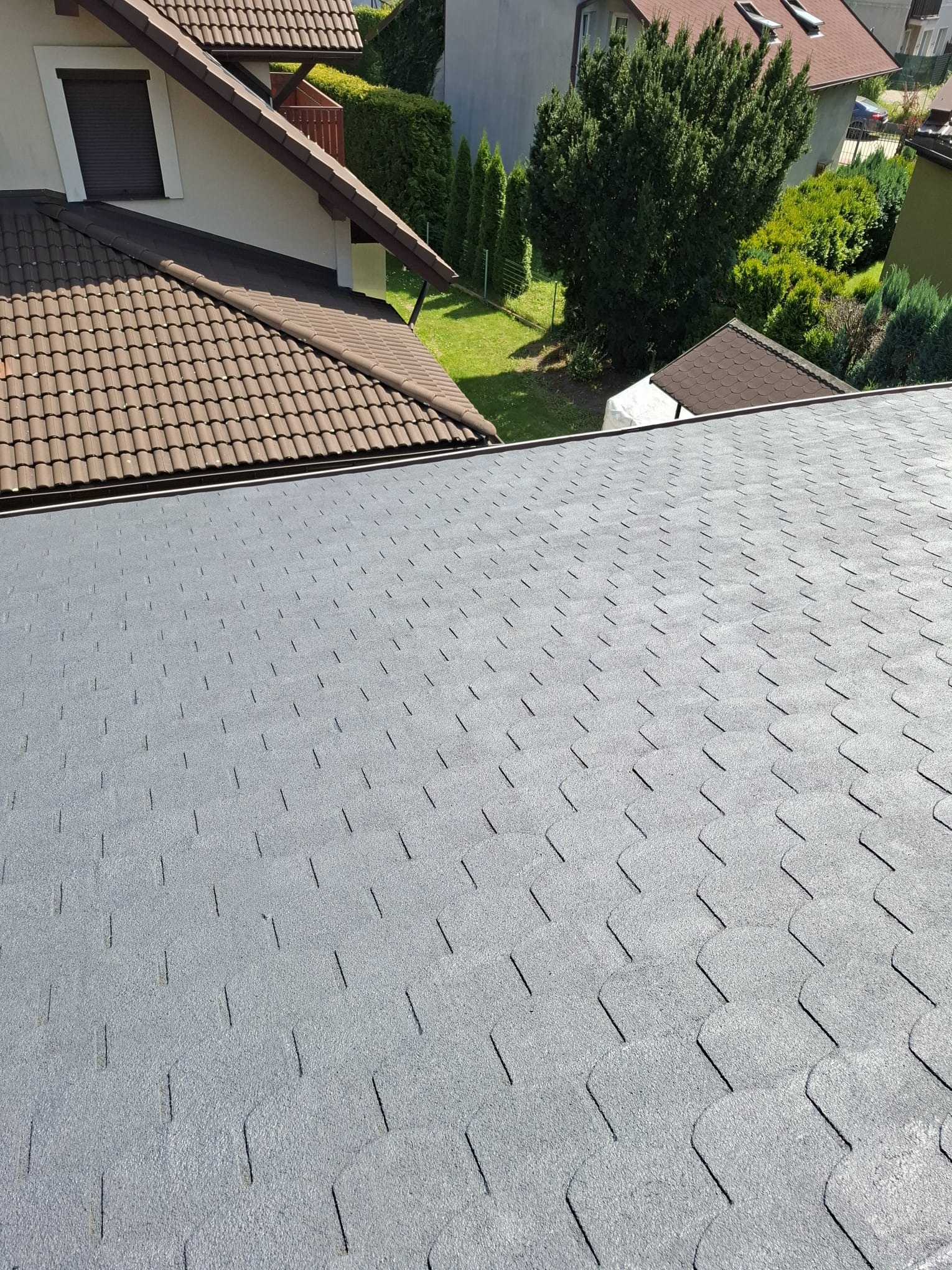 Szary dach pokryty gontem bitumicznym, widok z góry. W tle brązowy dach i zieleń drzew.
