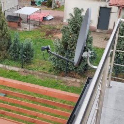 SAT-ANT - Szara antena satelitarna zamontowana na metalowym uchwycie, widoczna z balkonu na tle trawnika, ogrodzenia i budynku gospodarczego z szarą bramą.