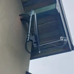 Zewnętrzna antena satelitarna zamontowana na ścianie budynku pod okapem dachu, widoczne kable i uchwyty mocujące.