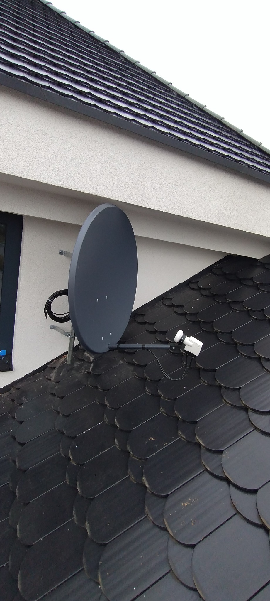 Szara antena satelitarna zamontowana na dachu krytym czarną dachówką ceramiczną, widok z dołu.