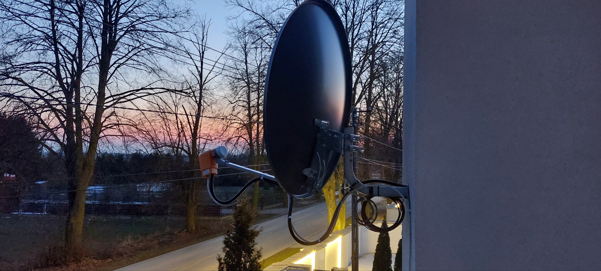 Czarna antena satelitarna zamontowana na ścianie budynku, z widokiem na drzewa i zachodzące słońce w tle.
