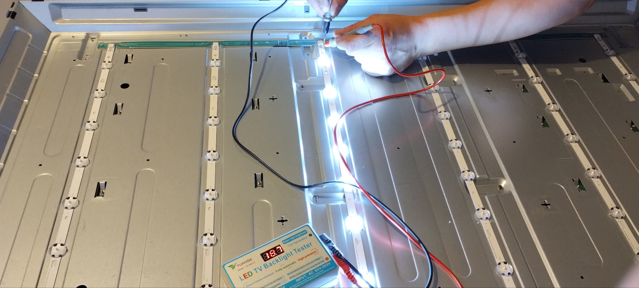 Testowanie podświetlenia LED telewizora za pomocą testera z wyświetlaczem 18.3V, widoczne paski LED z włączonymi diodami i ręka technika przykładająca sondę pomiarową.