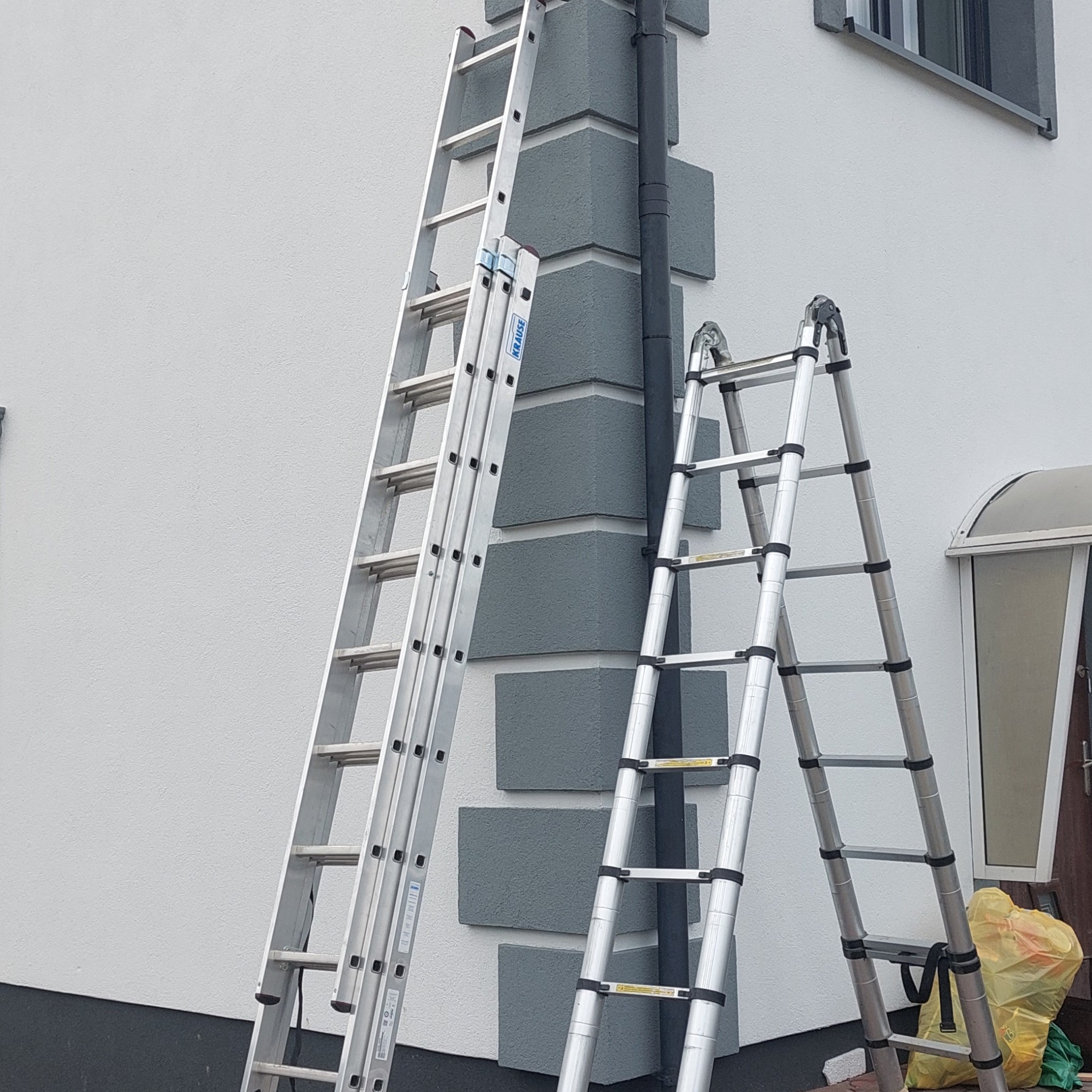 Dwie aluminiowe drabiny rozstawione przy elewacji domu z szarymi elementami dekoracyjnymi i rurą spustową.