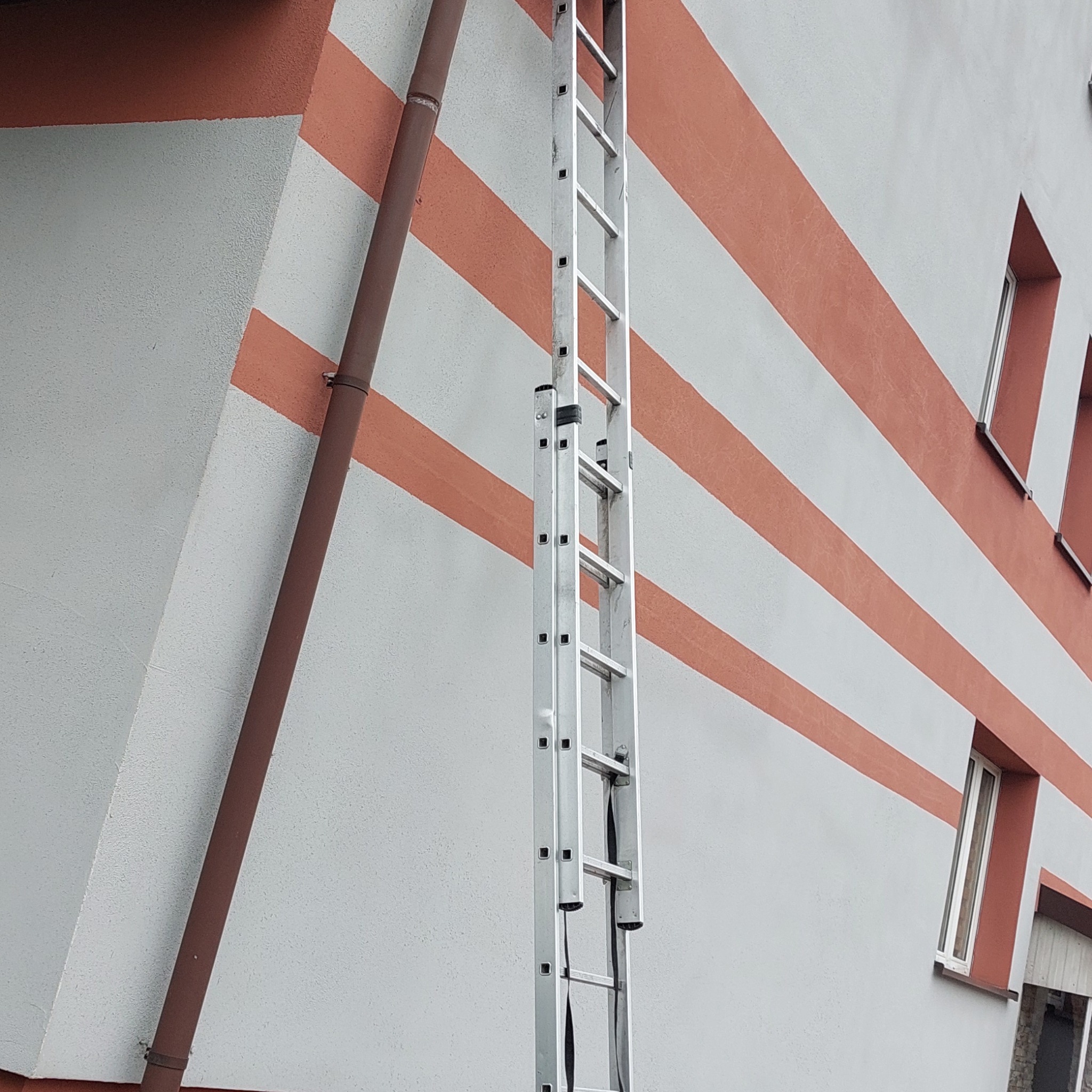 Aluminiowa drabina oparta o ścianę budynku z poziomymi, pomarańczowymi pasami i rynną spustową.