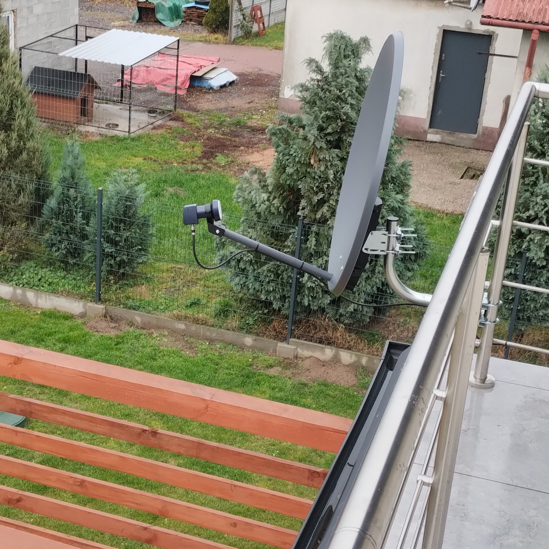 Szara antena satelitarna zamontowana na metalowym uchwycie, widoczna z balkonu na tle trawnika, ogrodzenia i budynku gospodarczego z szarą bramą.