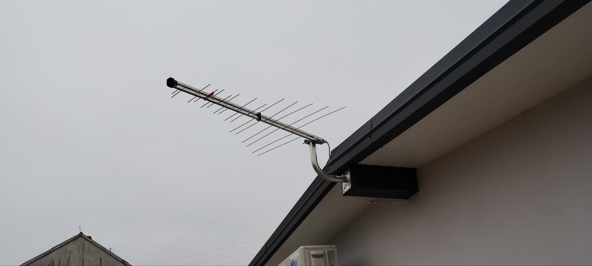 Antena telewizyjna Yagi-Uda zamontowana na ścianie domu pod okapem, widoczne elementy mocujące i skrzynka rozdzielcza.
