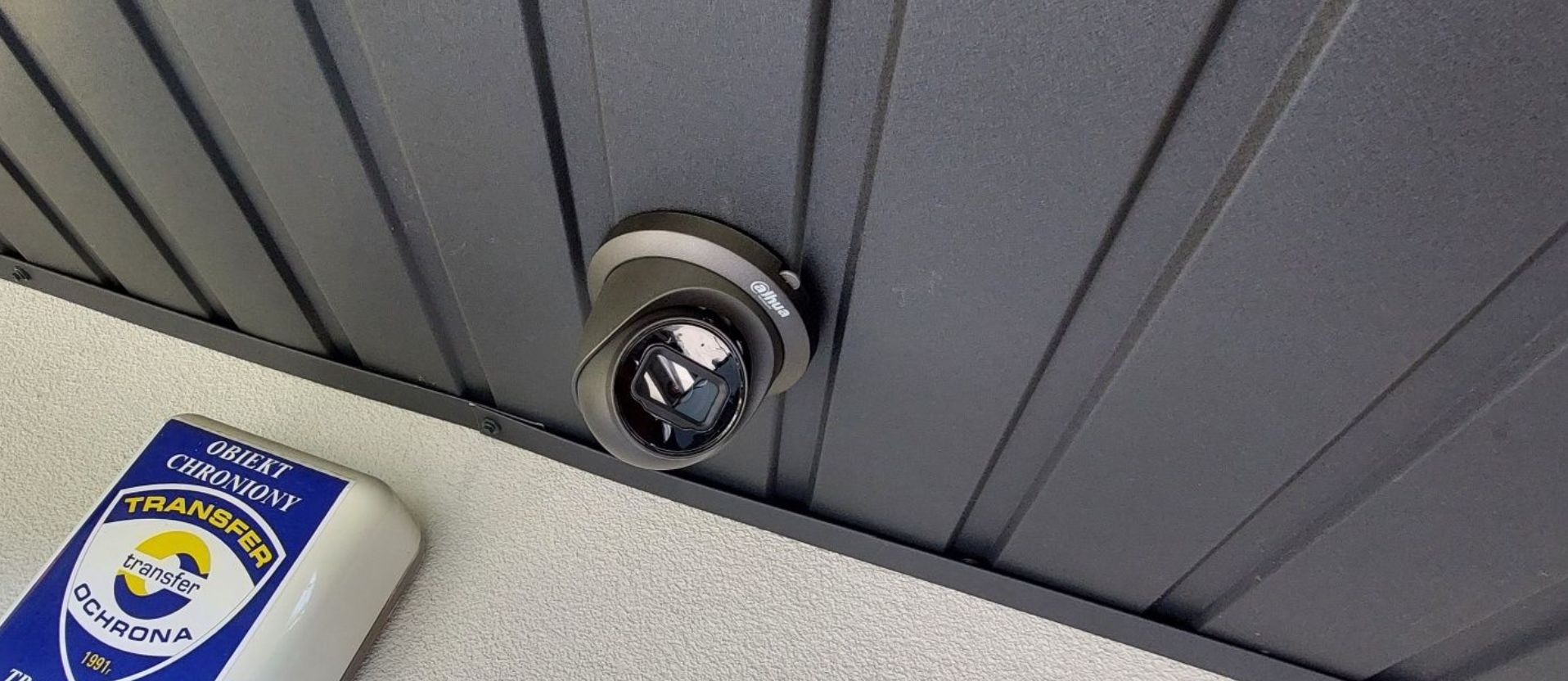 Kamera Dahua zamontowana pod szarym zadaszeniem, obok widoczna tabliczka informująca o obiekcie chronionym przez Transfer Ochrona.