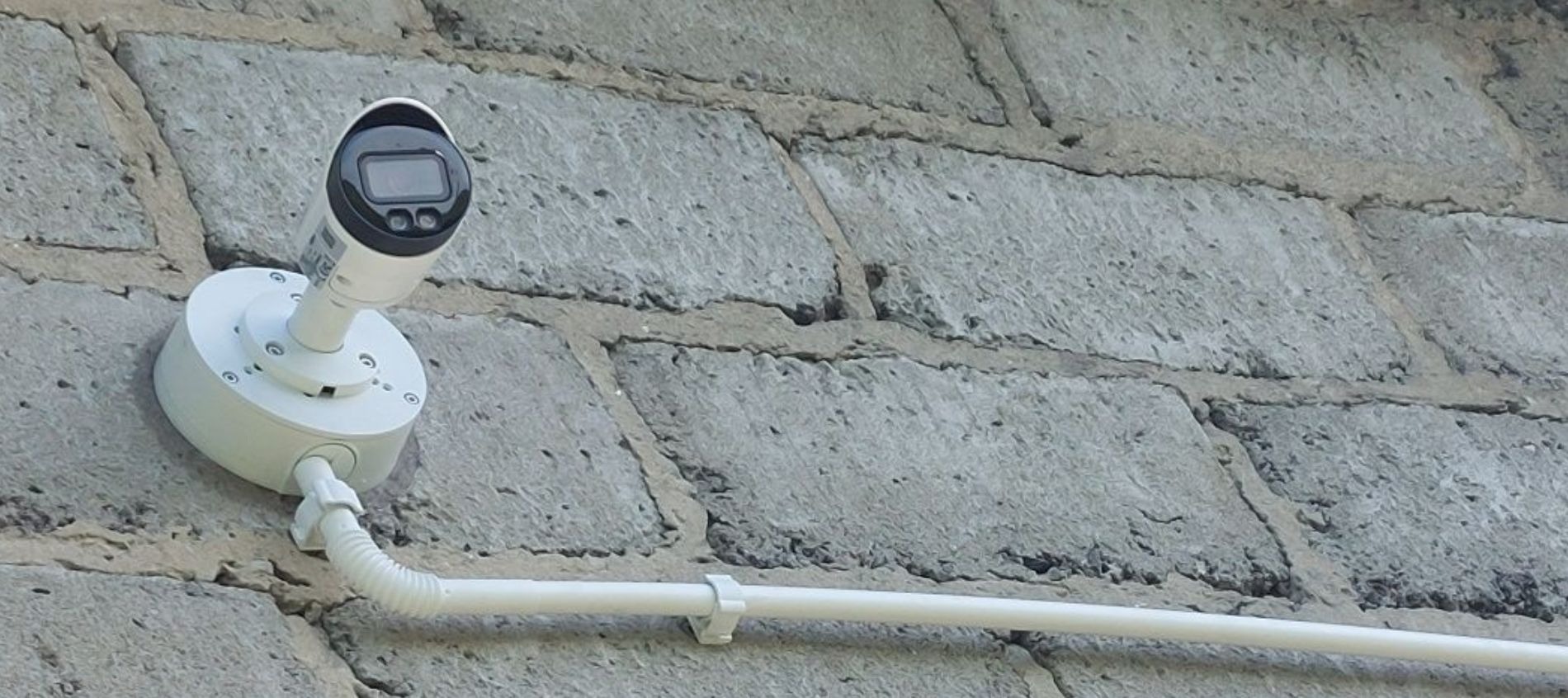 Kamera monitoringu z wyświetlaczem LCD zamontowana na szarej, betonowej ścianie. Widoczna biała puszka montażowa i peszel z kablem.