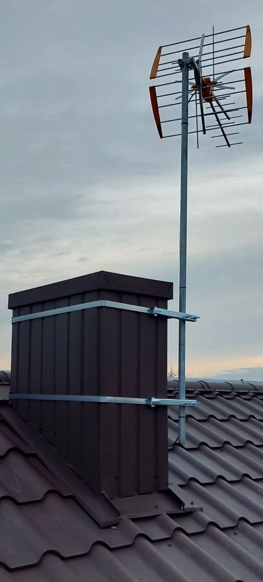 Antena telewizyjna Yagi-Uda zamontowana na metalowym maszcie, przymocowanym do brązowego komina na dachu z brązowej blachodachówki, na tle pochmurnego nieba.