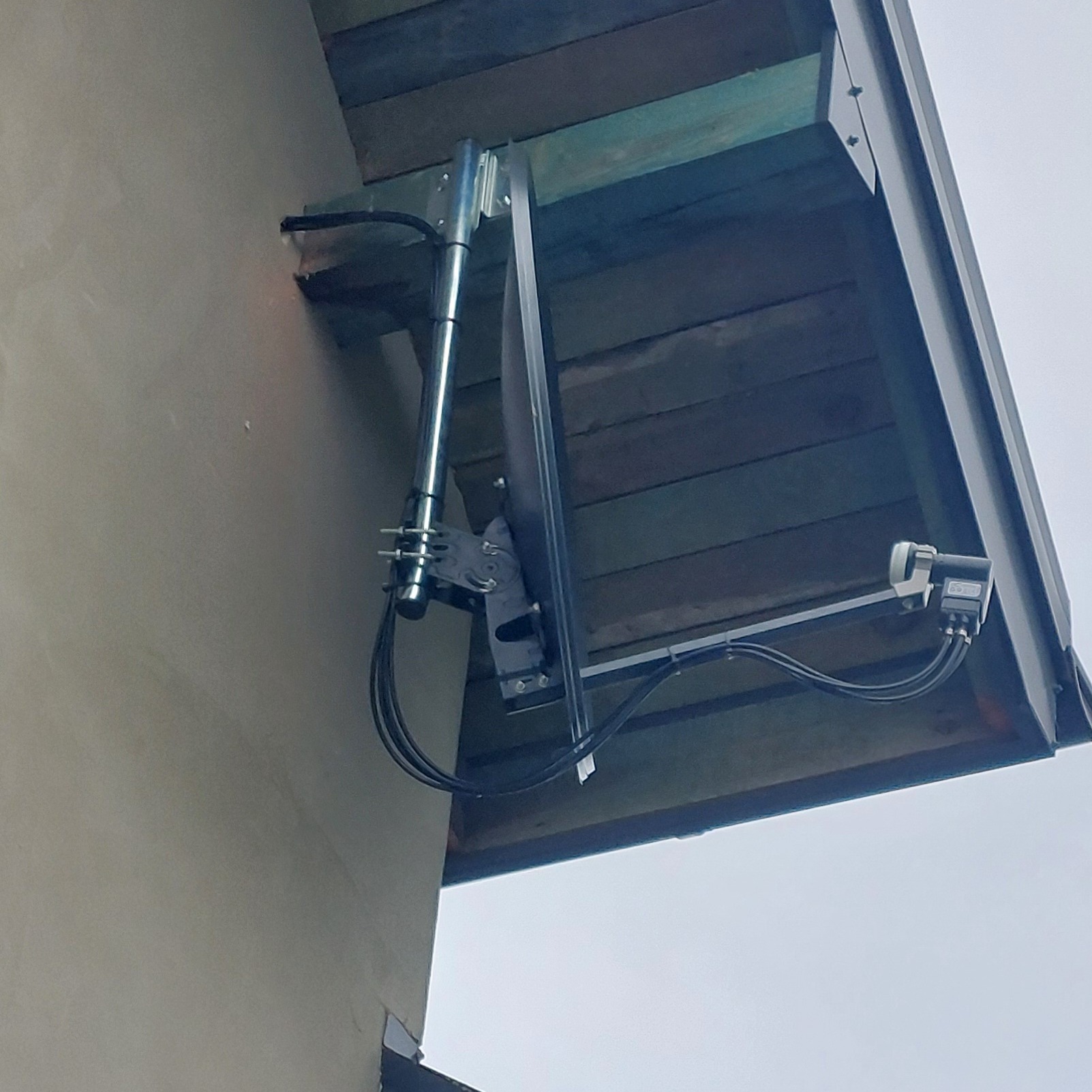 Zewnętrzna antena satelitarna zamontowana na ścianie budynku pod okapem dachu, widoczne kable i uchwyty mocujące.