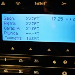 Panel sterowania systemu alarmowego Satel z wyświetlanymi temperaturami w różnych strefach domu: salon, piętro, garaż, piwnica i na zewnątrz. Widoczny fragment klawiatury numerycznej i symboli...