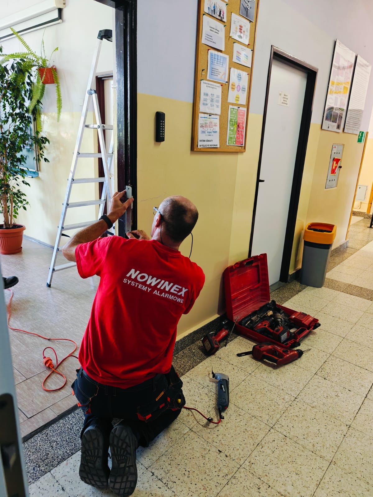 Instalator w czerwonej koszulce firmy NOWINEX SYSTEMY ALARMOWE klęczy przy futrynie drzwi, montując system kontroli dostępu. Obok rozłożona skrzynka z narzędziami, drabina oparta o ścianę,...