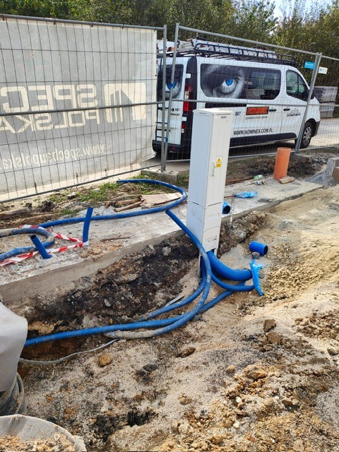 Biała skrzynka elektryczna z niebieskimi rurami karbowanymi w wykopie ziemnym, na tle ogrodzenia budowlanego i firmowego samochodu dostawczego z grafiką oka.