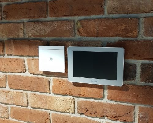 Panel sterowania alarmem i czujnik ruchu zamontowane na ścianie z czerwonej cegły.