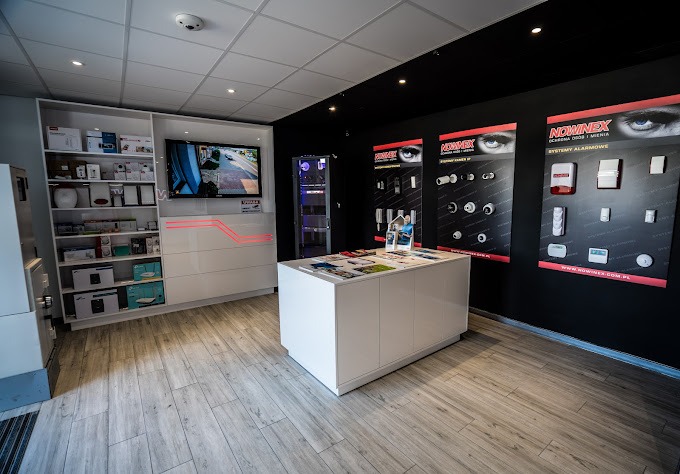Wnętrze sklepu z systemami alarmowymi i monitoringu, prezentacja produktów na ścianach i półkach, widoczny ekran z podglądem z kamery.
