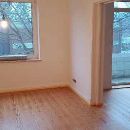 Leg-Bud - Pokój po odświeżeniu w Legnicy: świeżo pomalowane ściany na biało i odnowiona drewniana podłoga zabezpieczona taśmą malarską przy listwach.