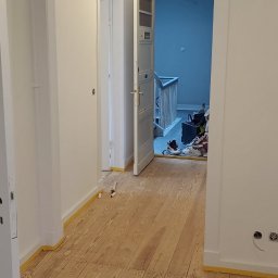 Leg-Bud - Korytarz w trakcie malowania: świeżo pomalowane białe ściany, drewniana podłoga zabezpieczona taśmą malarską, widoczne narzędzia i akcesoria malarskie w tle.