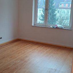 Leg-Bud - Pokój po malowaniu ścian z drewnianą podłogą zabezpieczoną taśmą malarską, widoczne niedokończone podłączenie elektryczne na suficie.