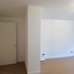 Leg-Bud - Pomalowane na biało wnętrze pokoju z beżową wykładziną, widoczne gniazdka elektryczne przy podłodze i biała szafka nocna z szufladą.