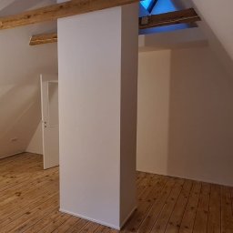 Leg-Bud - Poddasze po remoncie, świeżo pomalowane białe ściany, drewniana podłoga, widoczne drewniane belki stropowe, surowa żarówka na kablu.