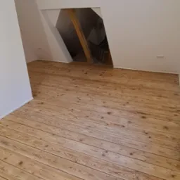 Poddasze z drewnianą podłogą z desek, widoczne drewniane belki stropowe i fragment schodów w tle.