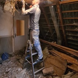Leg-Bud - Mężczyzna na drabinie usuwa fragment uszkodzonego sufitu, widoczne odsłonięte belki stropowe i gruz na podłodze.