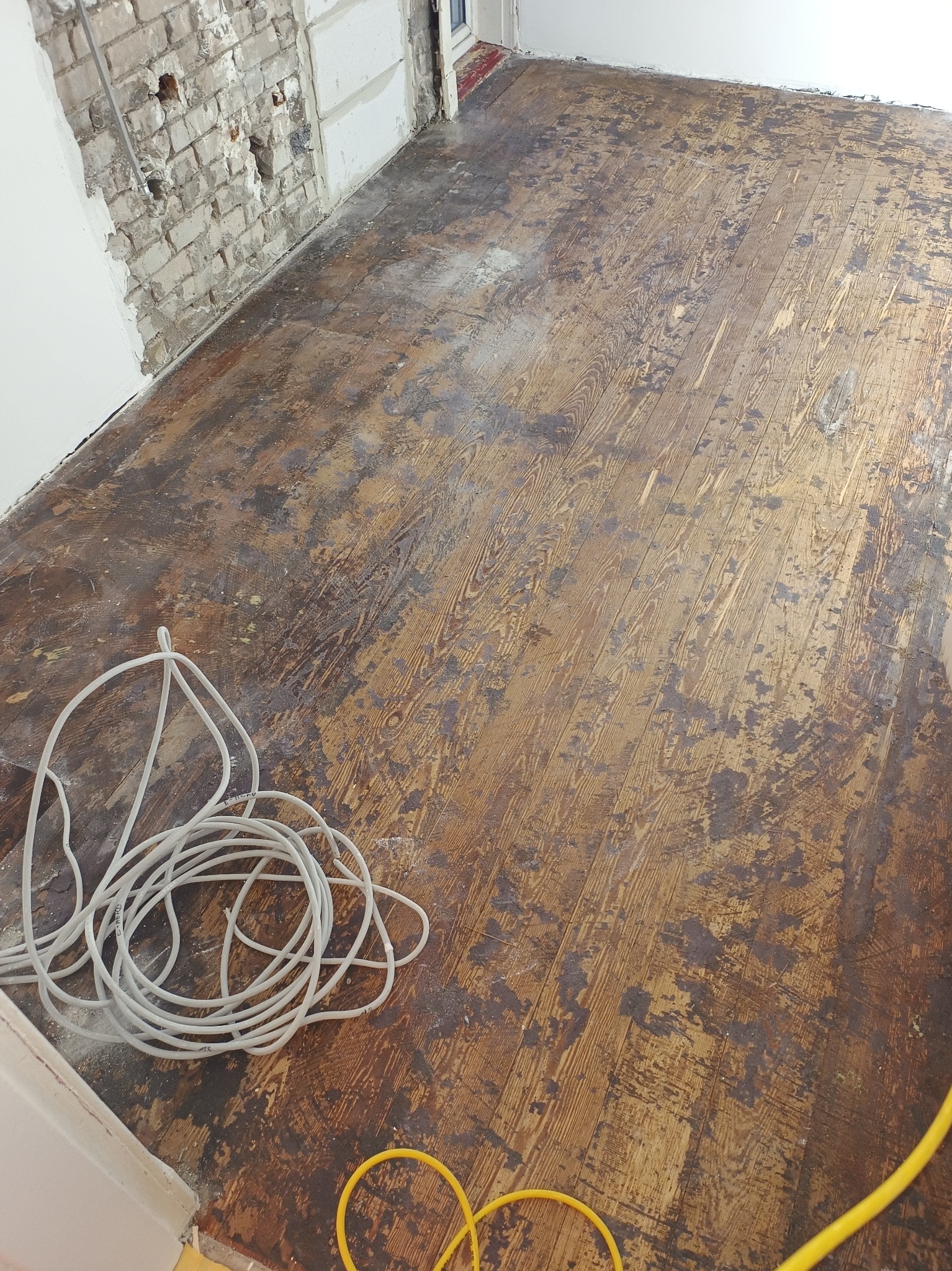 Podłoga z desek przed renowacją, z widocznymi śladami starej farby i kablami elektrycznymi w tle, fragment ściany z odsłoniętą cegłą.