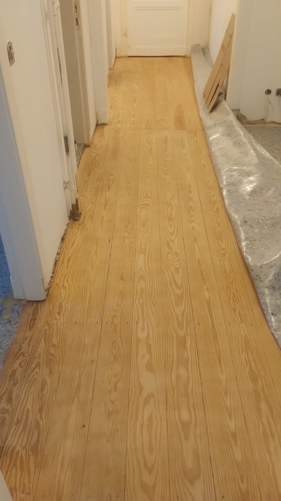 Odnowiona podłoga z jasnego drewna w korytarzu, zabezpieczona folią malarską przy ścianie, widoczne ślady po taśmie malarskiej przy listwach przypodłogowych.